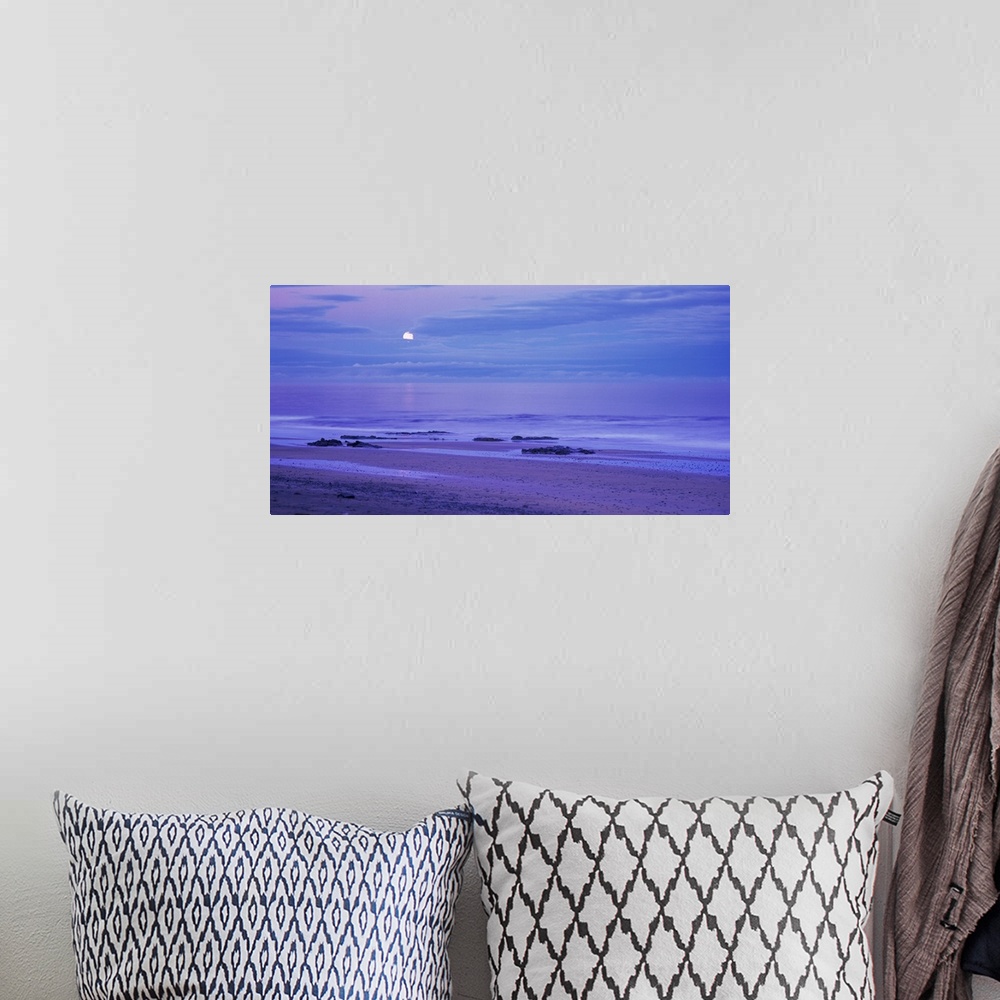 A bohemian room featuring The moon is seen slightly behind a cluster of clouds that hang over the ocean water and photograp...