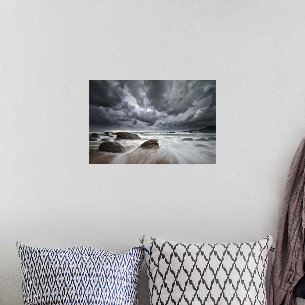 A bohemian room featuring Dramatic clouds hang over a rocky beach with the tide rushing out to sea.