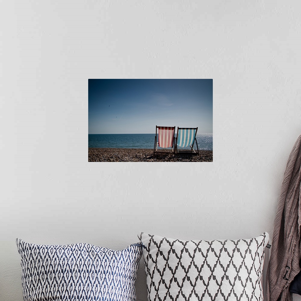 A bohemian room featuring Red and blue striped deckchair and sit facing out to blue sea. Pebble beach and clear blue sky.