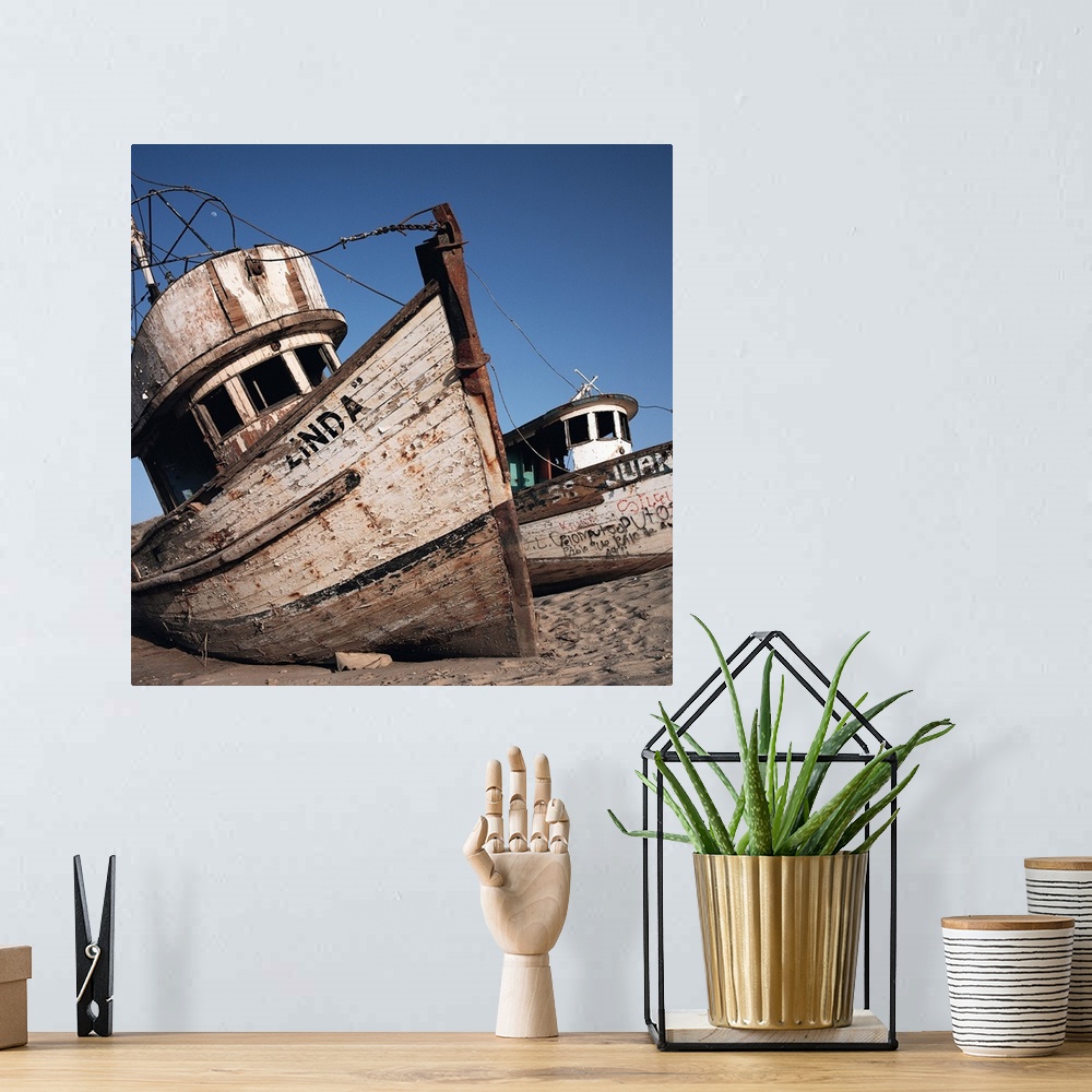 A bohemian room featuring old neglected boats sit beached on sand under a blue sky