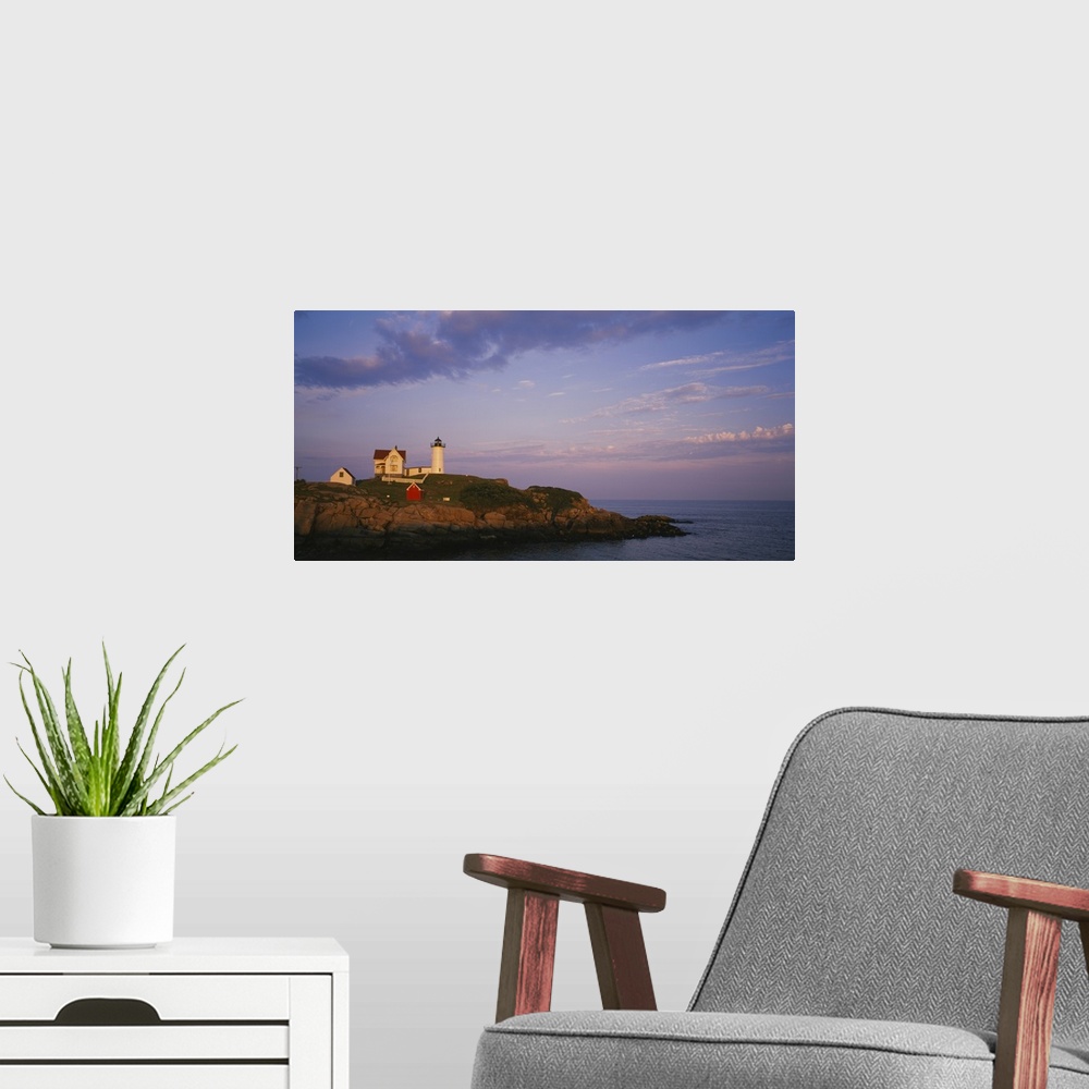 A modern room featuring Cape Neddick Lighthouse perched on the top of a green and rocky cliff with the warm sunset reflec...