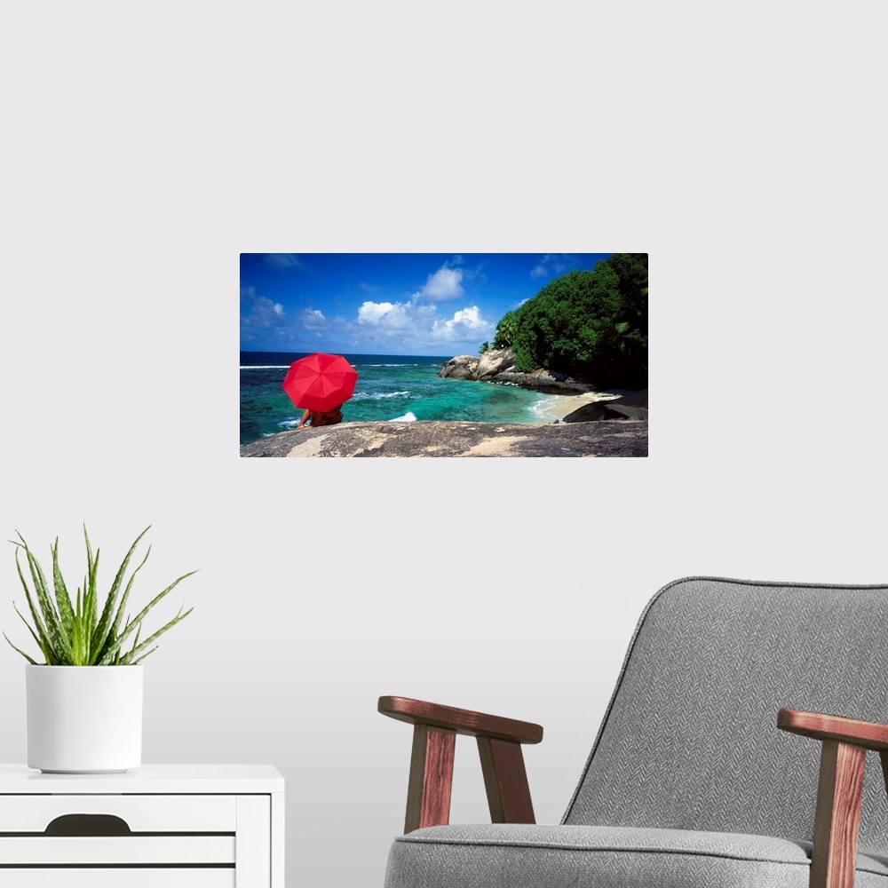 A modern room featuring A woman holding a red umbrella sits on a large rock and looks out over the ocean with the coast j...