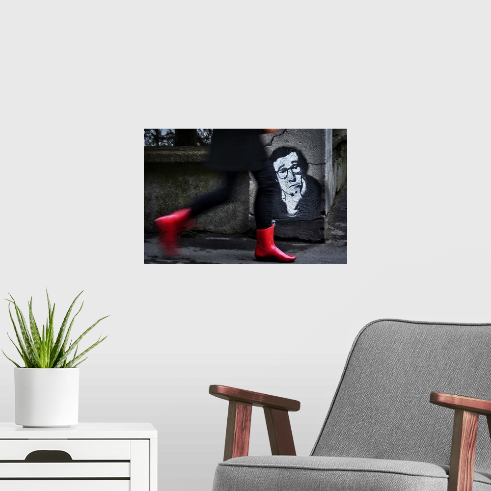 A modern room featuring A woman in red boots walks quickly down a street past a stenciled image of Woody Allen on a concr...