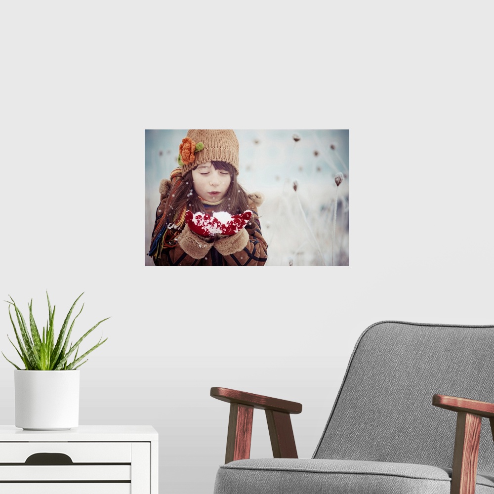 A modern room featuring A young girl in winter clothes blows on a handful of snow.