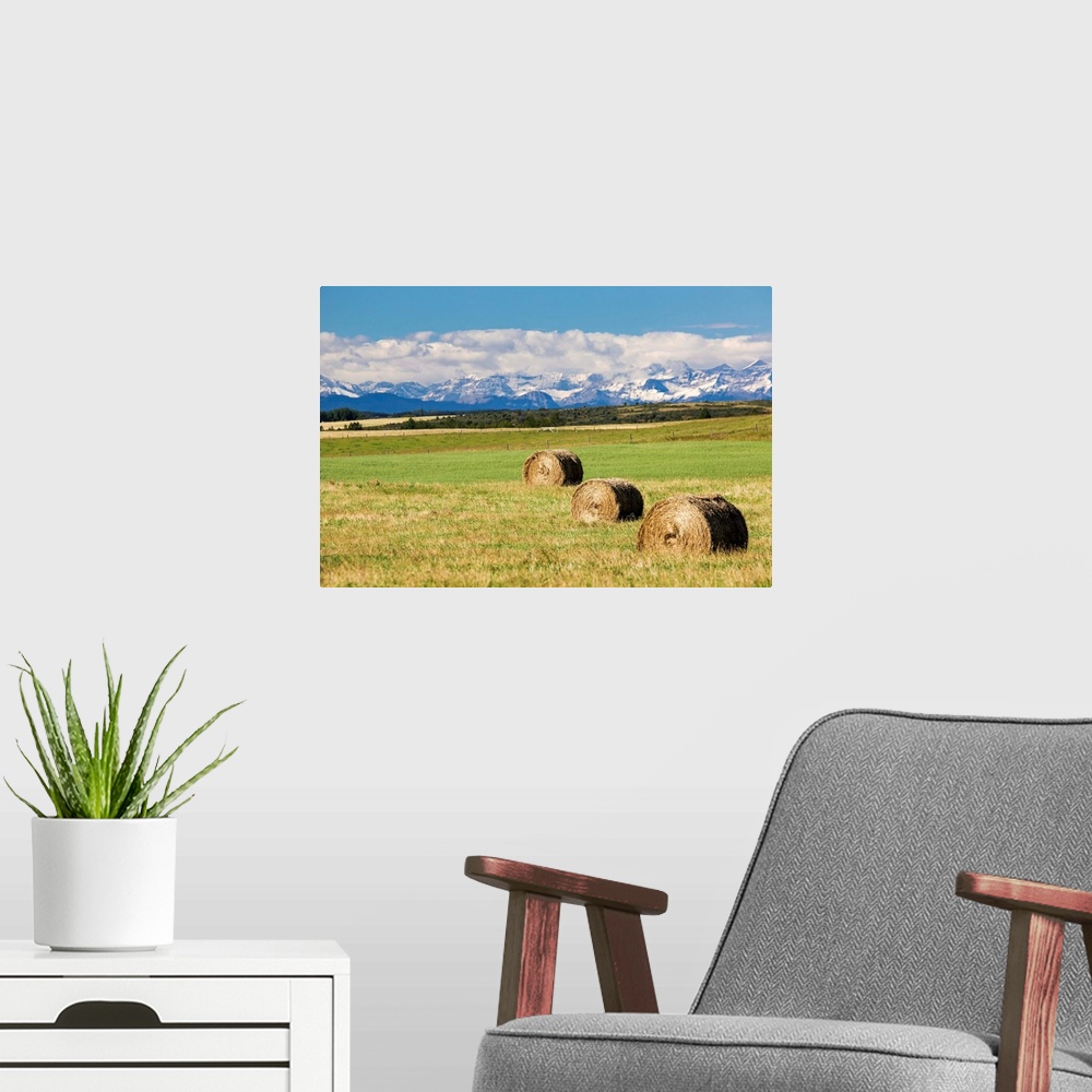 A modern room featuring Three hay bales in a field with mountains in the background slightly snow covered and cloudy with...