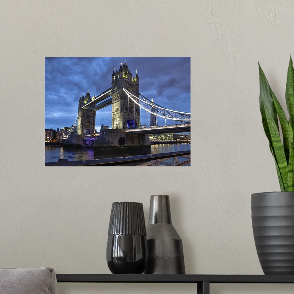 A modern room featuring Tower Bridge illuminated at dusk and reflected in the tranquil water of the River Thames; London,...
