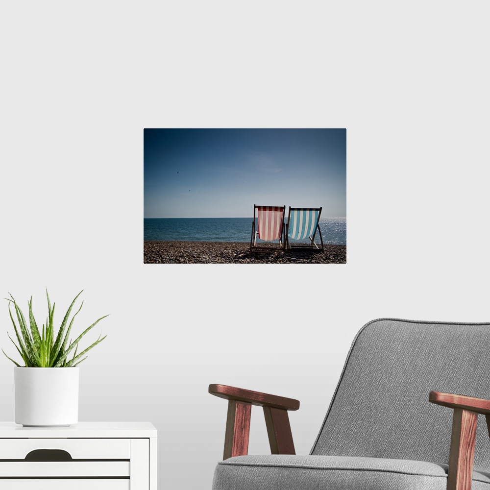A modern room featuring Red and blue striped deckchair and sit facing out to blue sea. Pebble beach and clear blue sky.