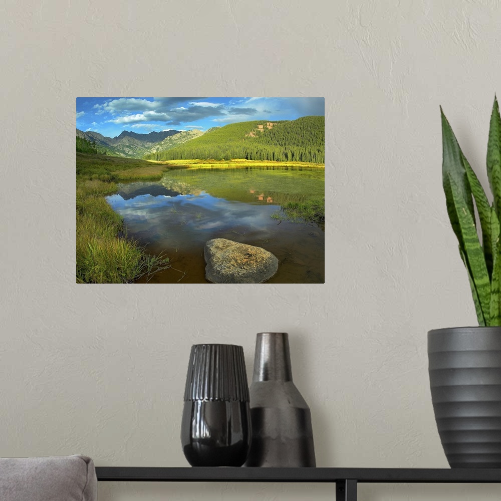 A modern room featuring This decorative wall art is a landscape photograph of a meadow, tree covered hills, and a mountai...