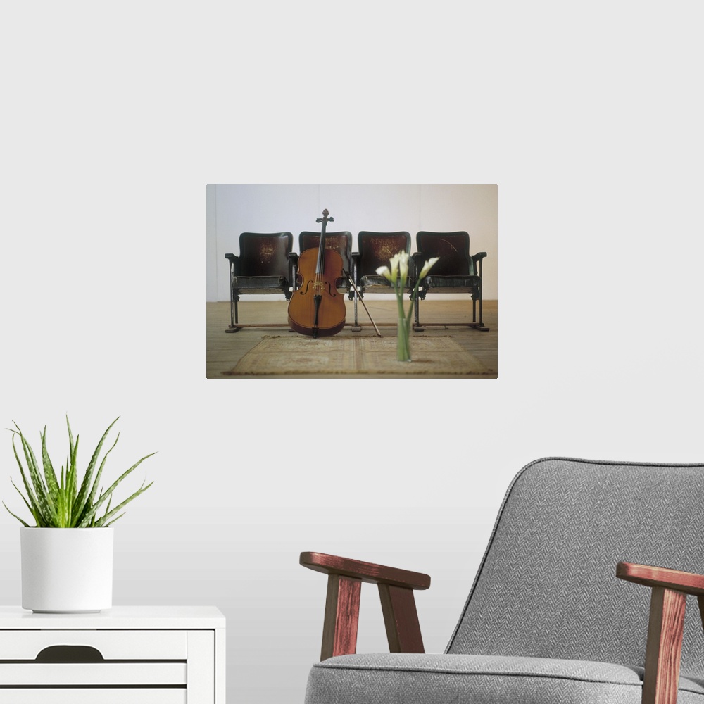 A modern room featuring A cello stands on the floor and leans against a row of four seats. A vase of calla lilies sits on...