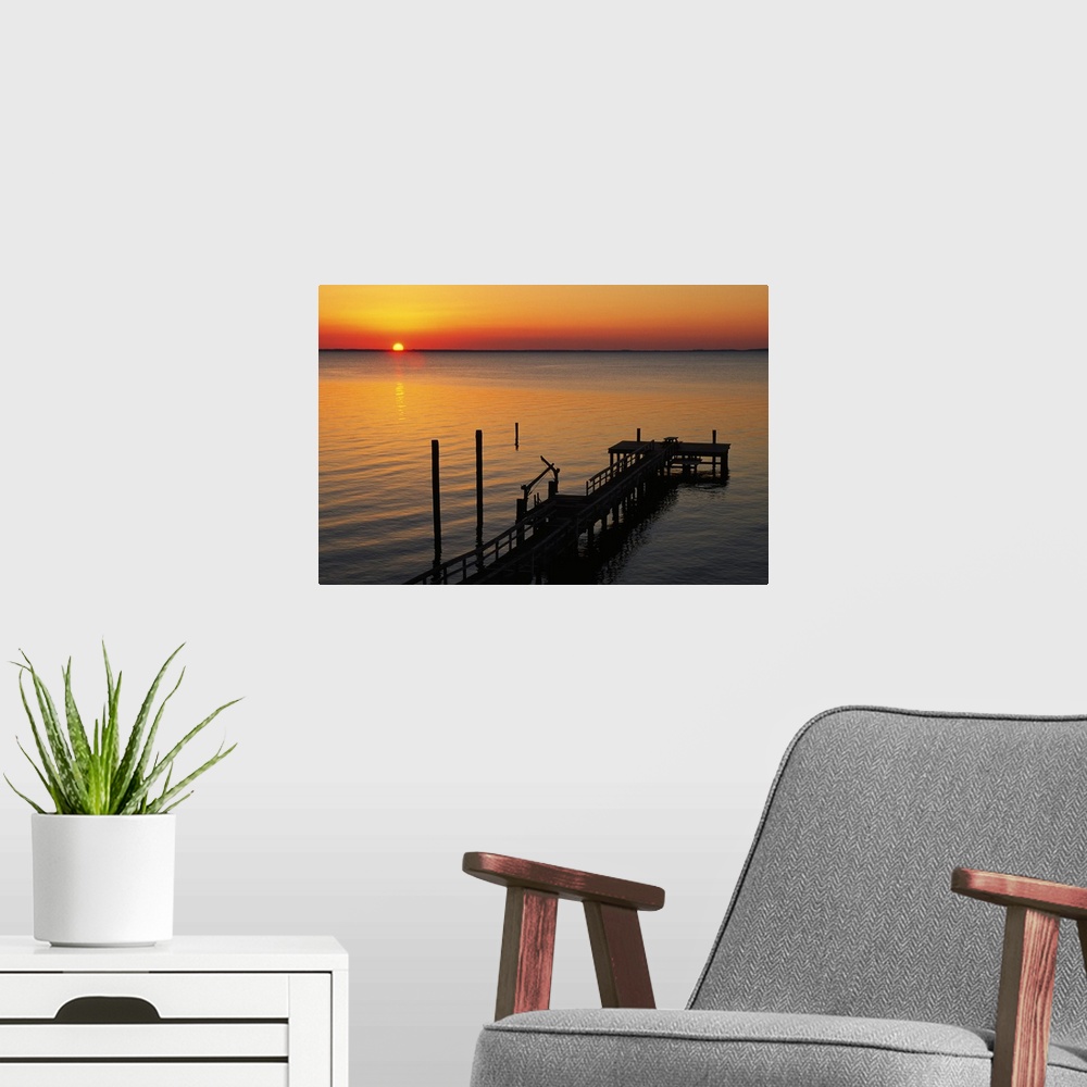 A modern room featuring A long pier is photographed reaching out into the ocean with the sun setting just on the horizon.