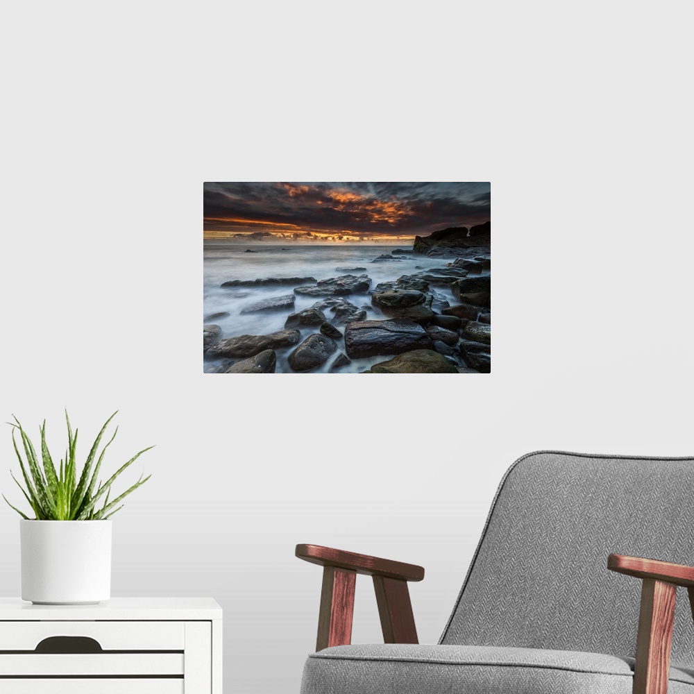 A modern room featuring Dramatic sky filled with dark clouds illuminated by the setting sun, with a rocky coastline in th...