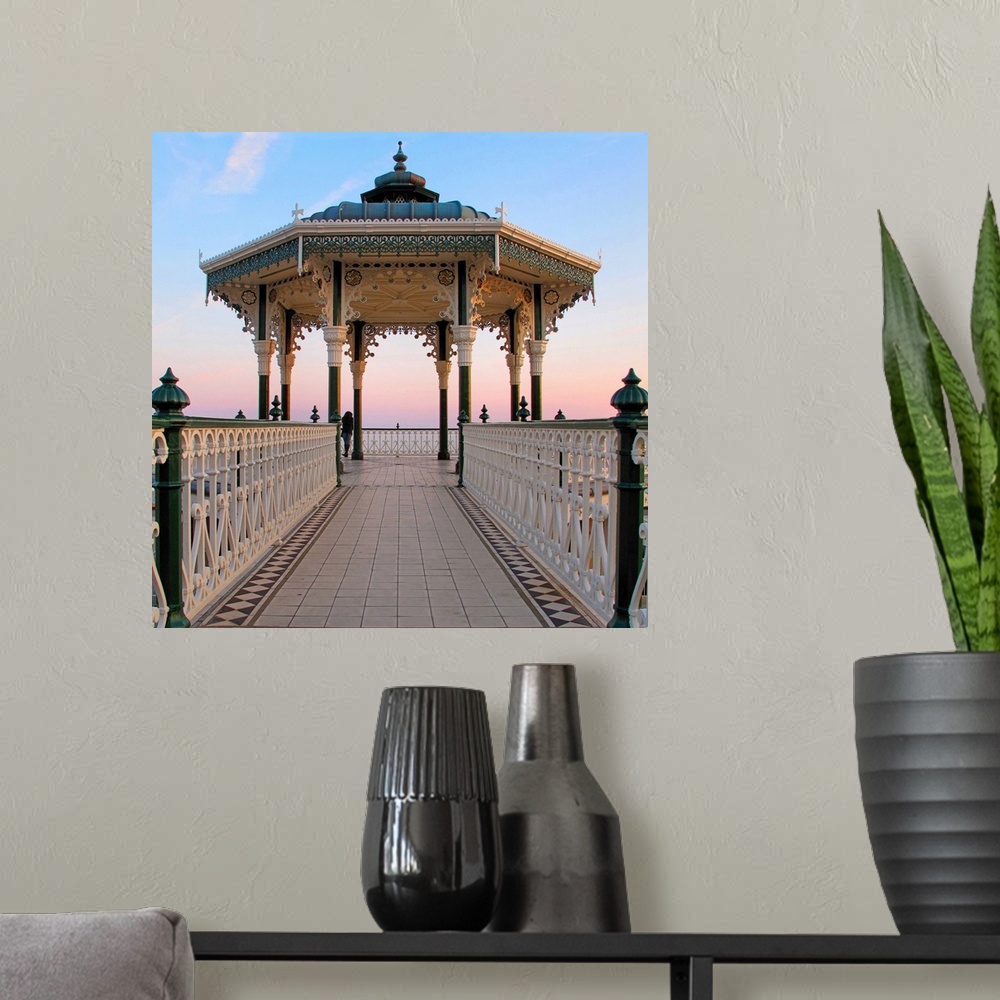 A modern room featuring A lone figure and an almost symmetrical shot of an ornate seaside bandstand. The sky is awash wit...