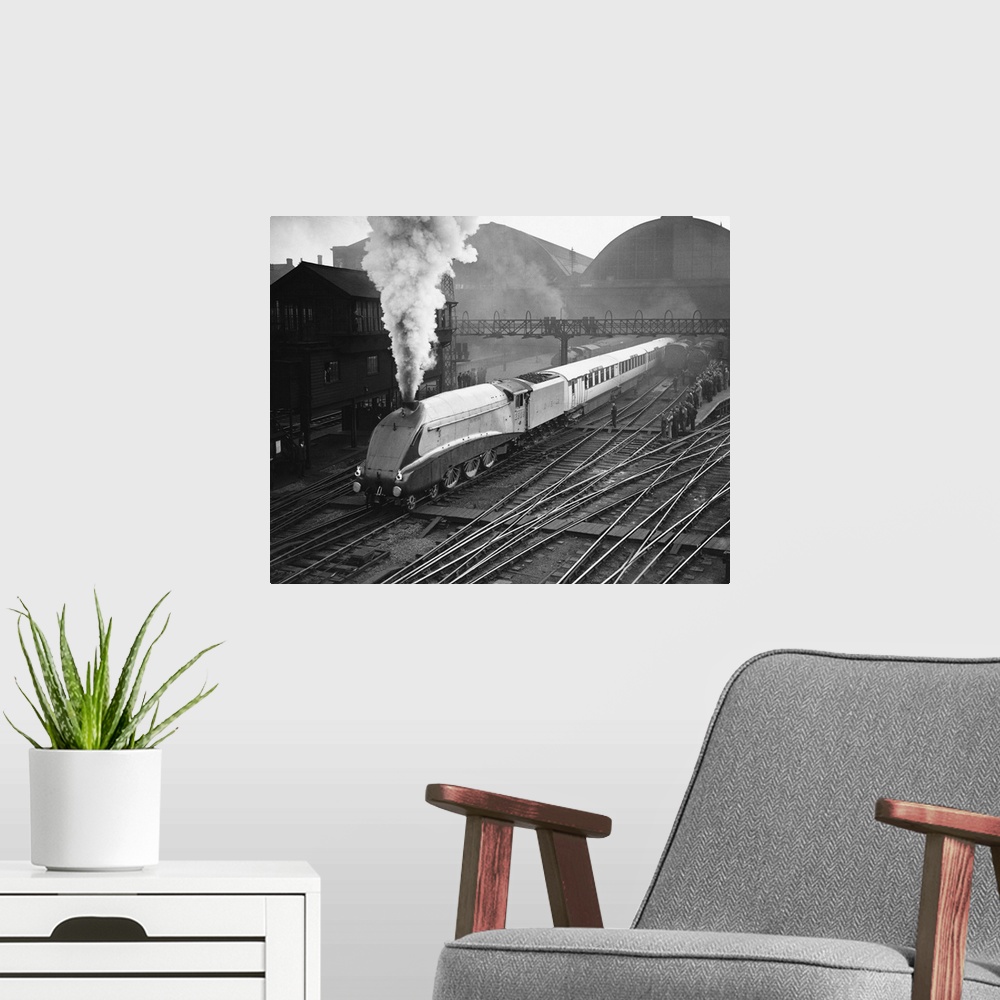 A modern room featuring The gleaming Silver Link emits a jet of steam as it passes a platform crowded with onlookers.