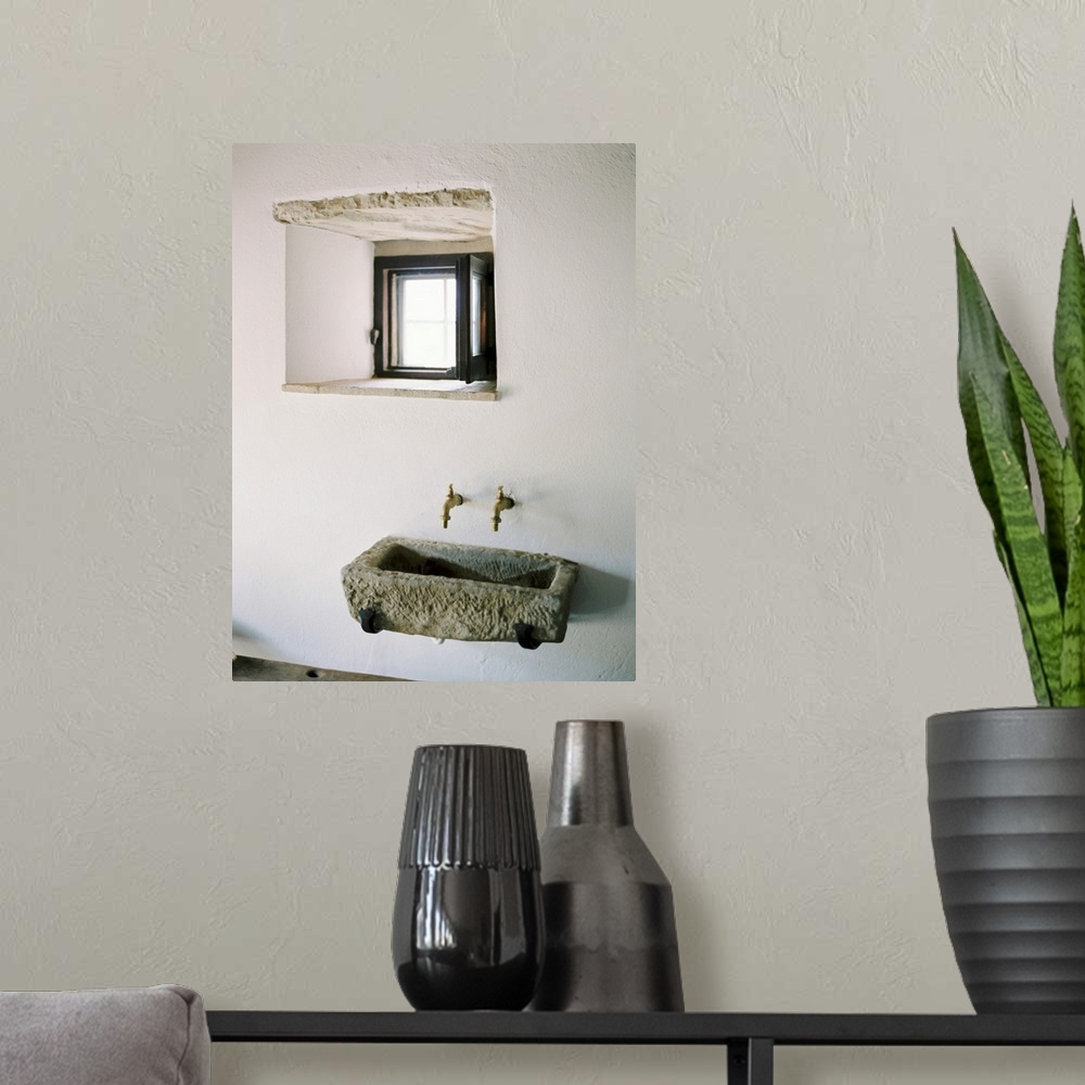 A modern room featuring A photograph of a rustic stone wall basin underneath a small window in a white stucco mediterrane...