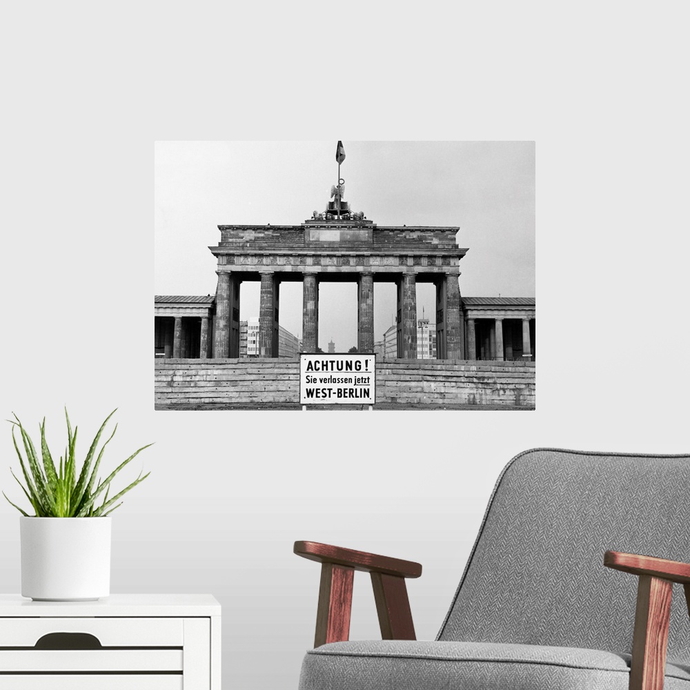 A modern room featuring The Brandenburg Gate in East Berlin. In front, a sign in German warns of the impending border bet...