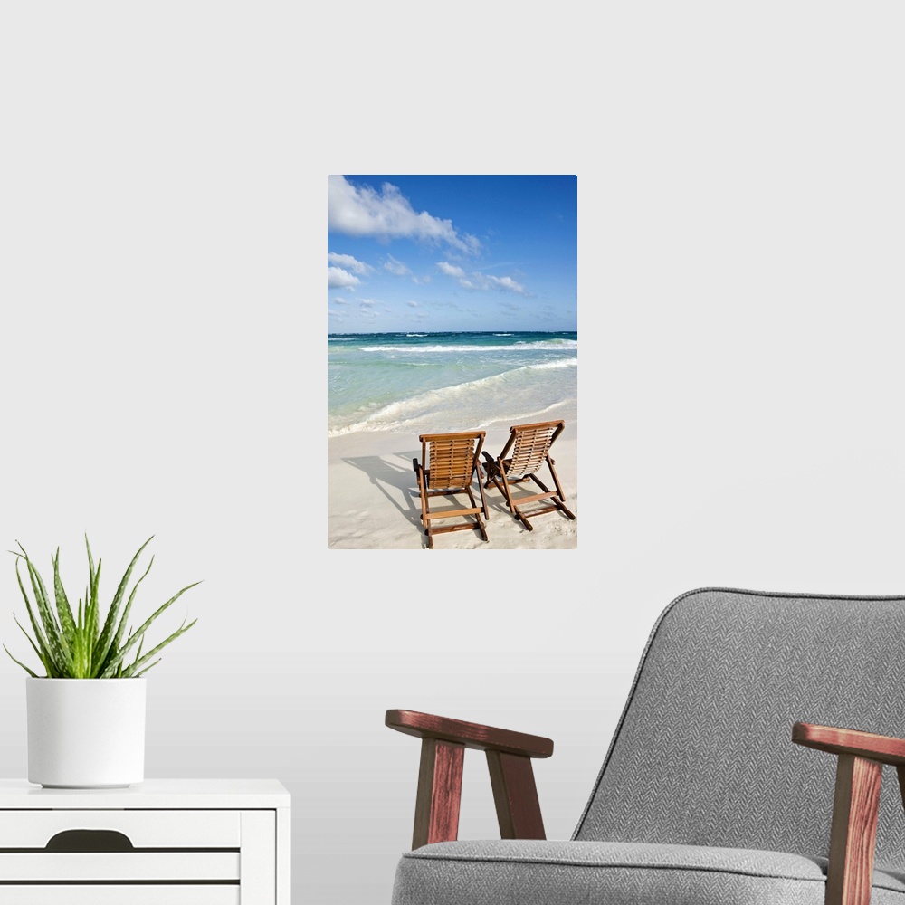 A modern room featuring Portrait photograph on a large wall hanging of two wooden lounge chairs on the beach, facing clea...