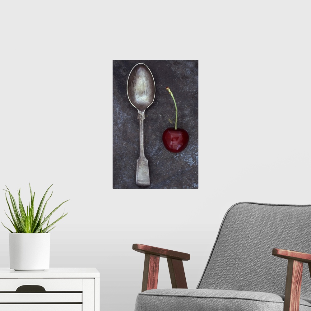 A modern room featuring Antique tarnished silver teaspoon lying next to single dark red cherry with stalk on tarnished metal