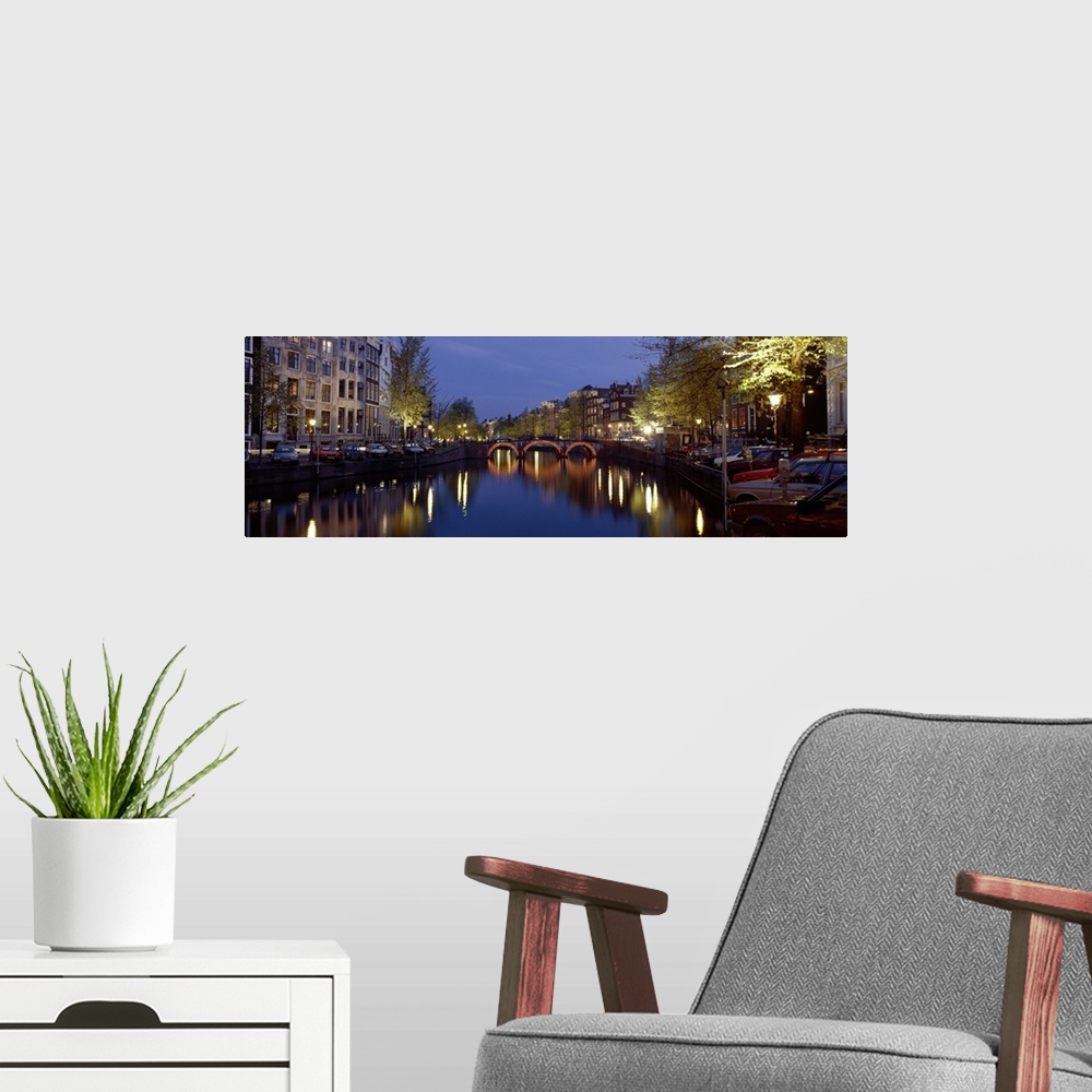 A modern room featuring A wide angle view of a canal in Amsterdam with buildings and street lights illuminated on either ...