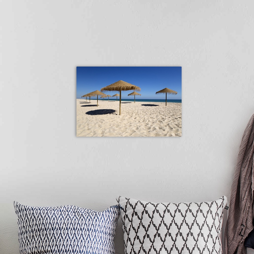 A bohemian room featuring Straw umbrellas on empty white sand beach with clear sea behind, Ilha do Farol, Culatra Barrier I...