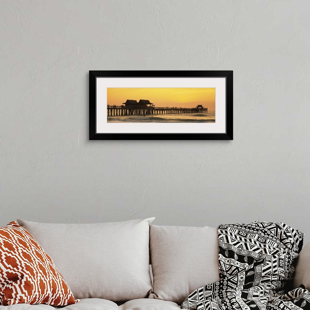 A bohemian room featuring A silhouetted wooden wharf with small buildings stretches out into the ocean at sunset. A couple ...