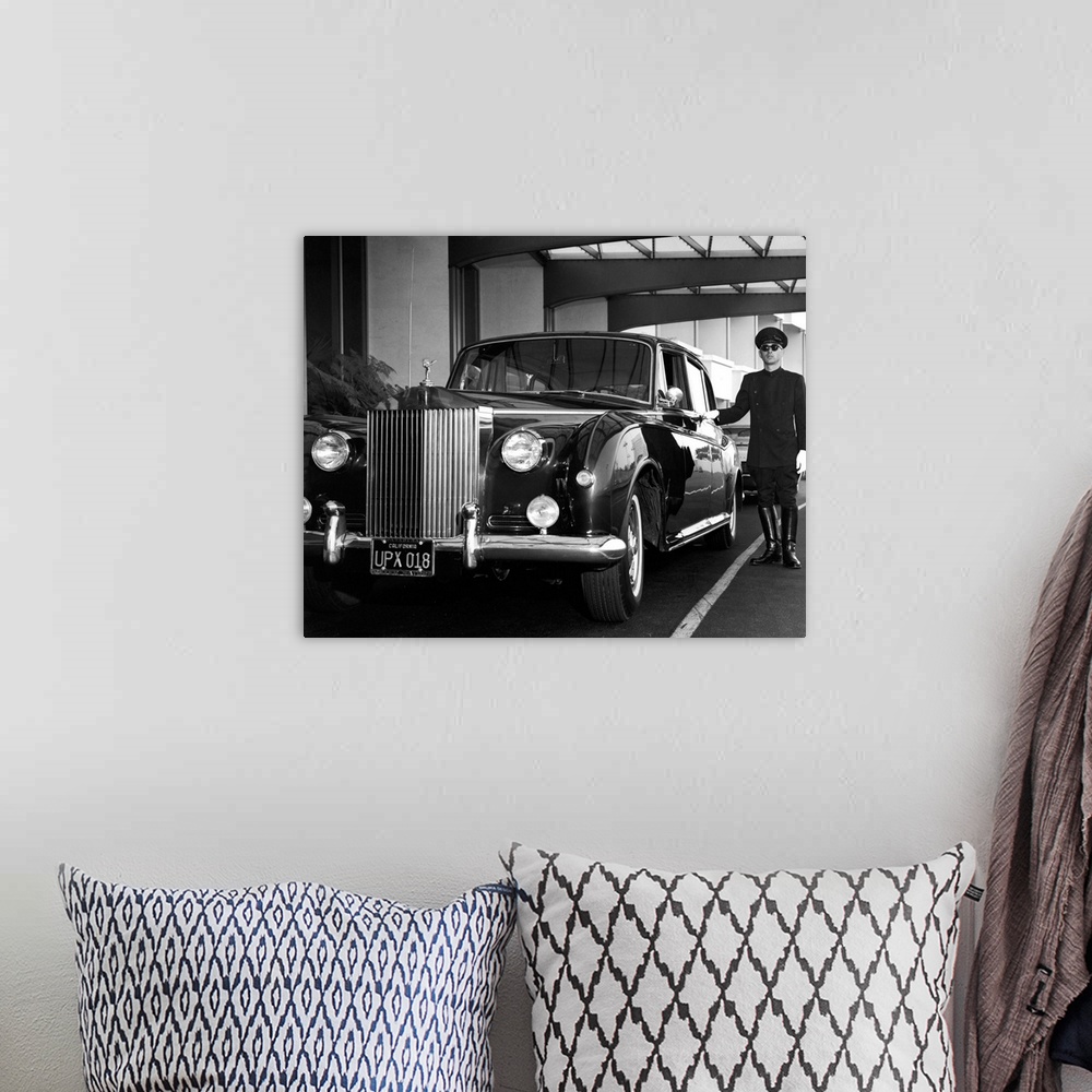 A bohemian room featuring A uniformed chauffeur waits beside a Rolls-Royce Phantom V, California, circa 1959