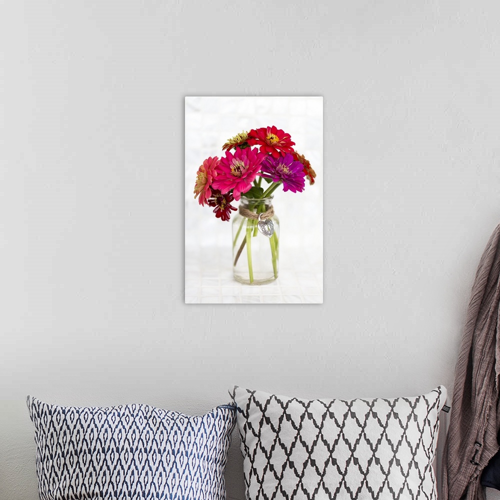 A bohemian room featuring A variety of colored Zinnia flowers in a simple glass vase with a decorative heart pendant held i...