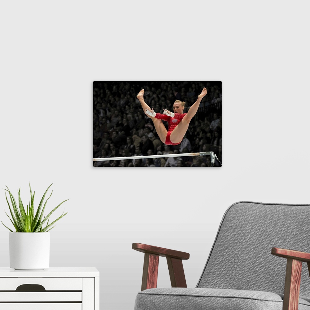 A modern room featuring A female gymnast flies over the high bar with her legs raised up, arms outstretched ready to grab...