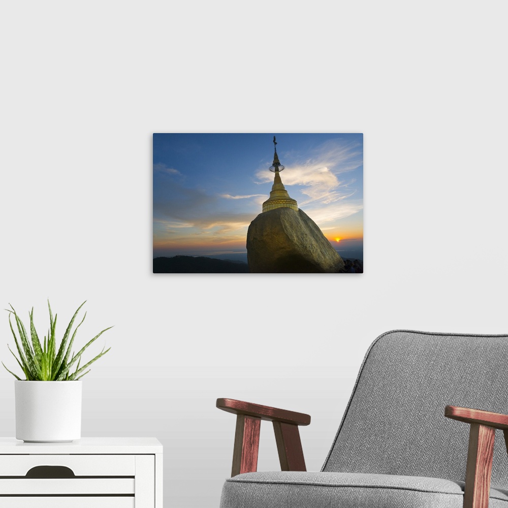 A modern room featuring Kyaiktiyo Pagoda at sunset, a small pagoda built on the top of a granite boulder covered with gol...