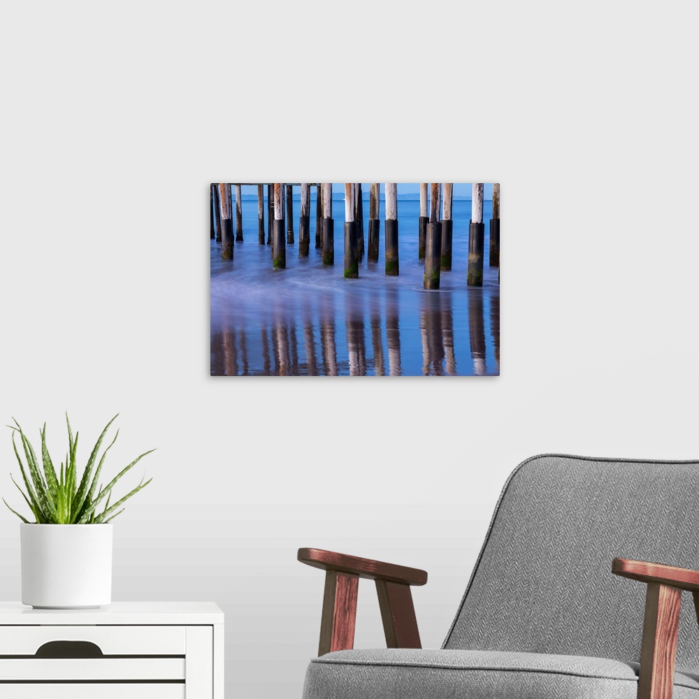 A modern room featuring Photograph of the bottom of Ventura Pier pilings reflecting into the ocean water with mountains i...