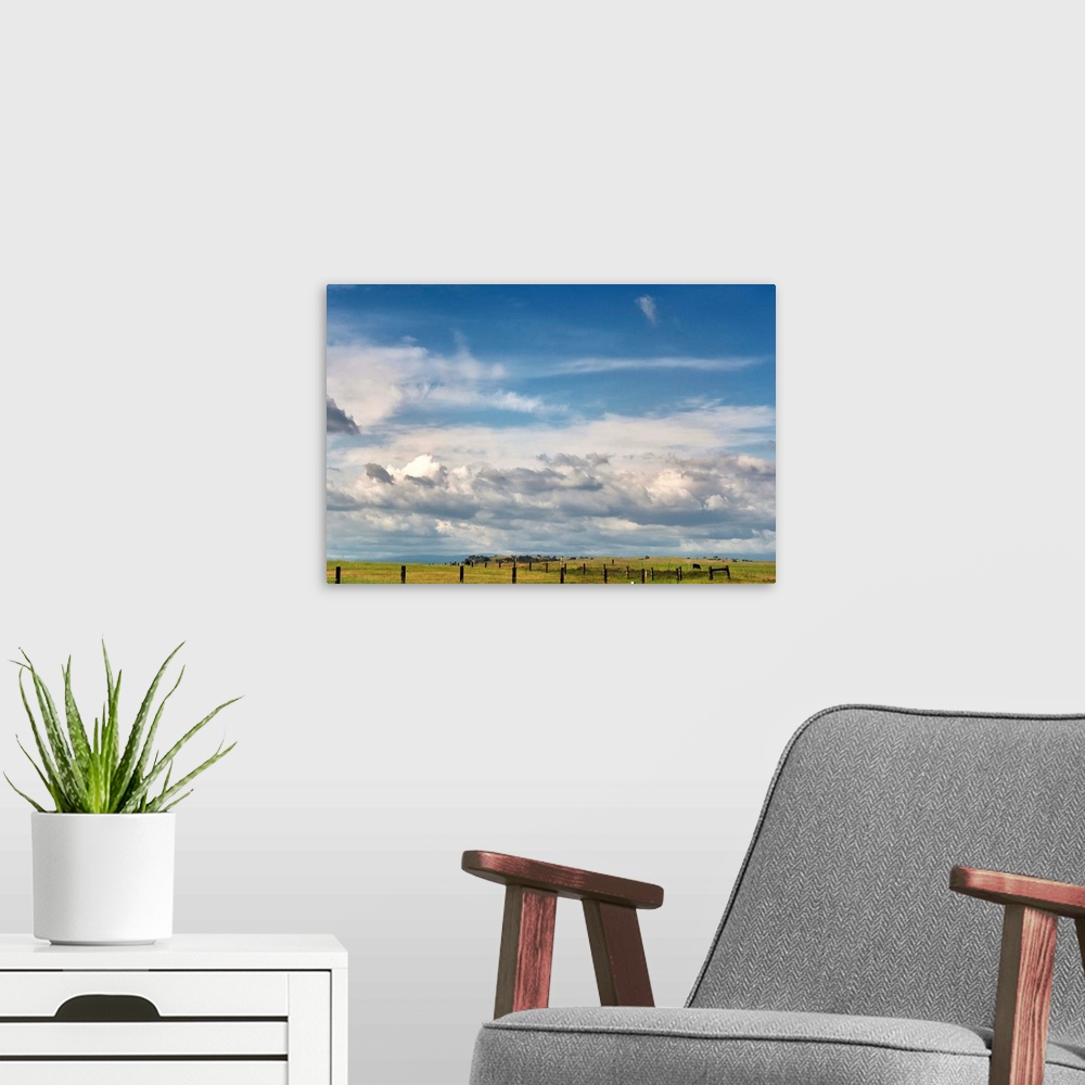 A modern room featuring Big blue sky with cumulus clouds fills most of image with slim view of grassland and fences of we...