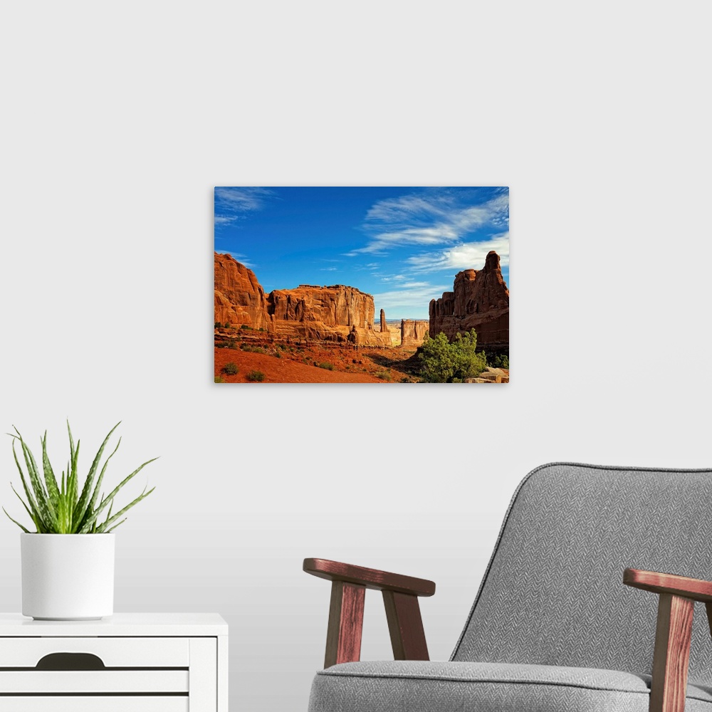 A modern room featuring Massive rock formations rise out of the desert floor with sheer cliff faces. Several shrubs grow ...