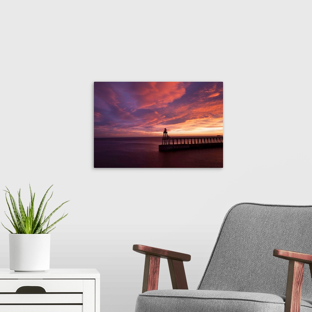 A modern room featuring A dramatic peach, orange and red sunset over the lighthouse and pier at Whitby harbour, North Yor...
