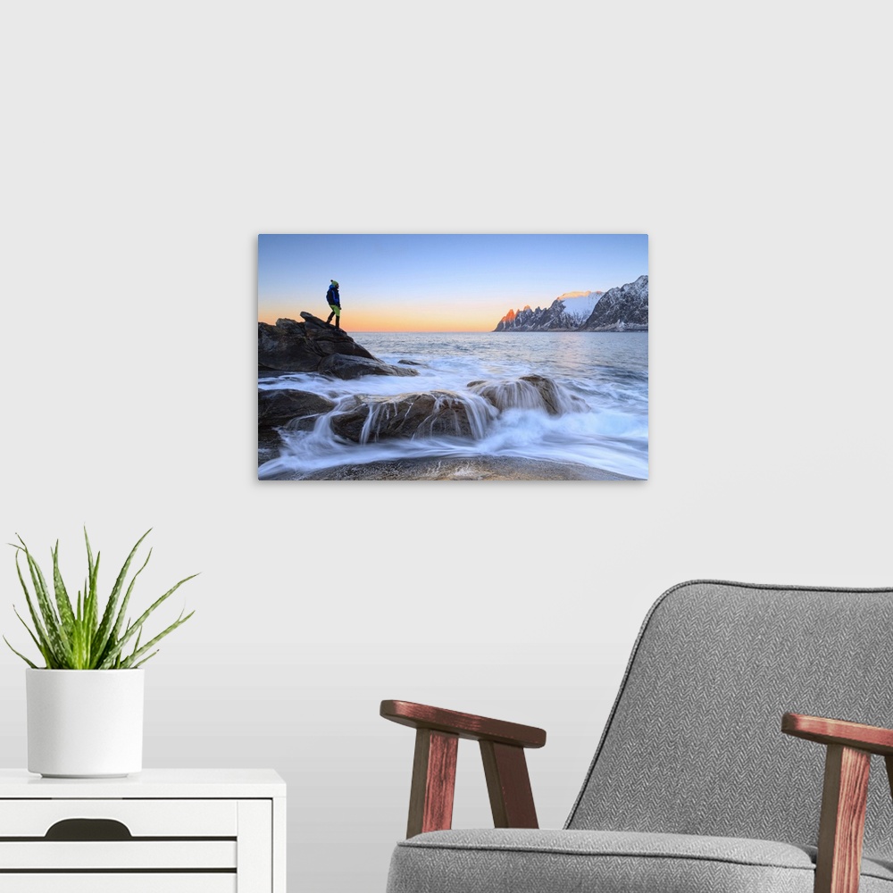 A modern room featuring Hiker admires the waves of the icy sea crashing on the rocky cliffs at dawn, Tungeneset, Senja, T...