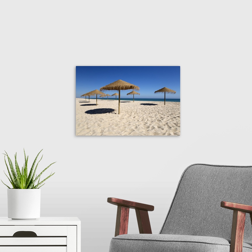 A modern room featuring Straw umbrellas on empty white sand beach with clear sea behind, Ilha do Farol, Culatra Barrier I...