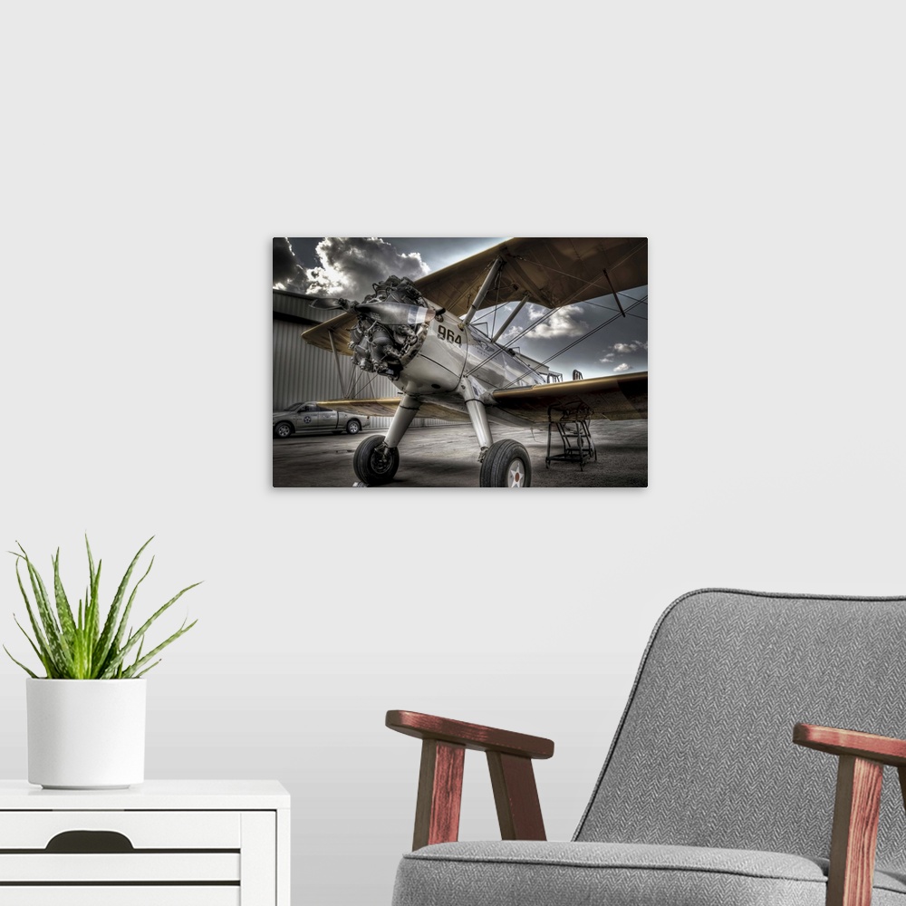 A modern room featuring A HDR photograph of a vintage air craft parked on the tarmac outside a hanger and a sky full of l...