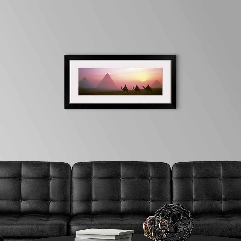 A modern room featuring Panoramic photograph of three camel riders at dusk with pyramids in the background.  The sun is s...