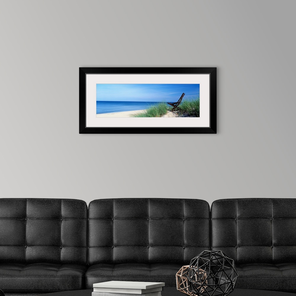A modern room featuring This wall art is a panoramic landscape photograph of a sandy beach with a chair in the dunes over...