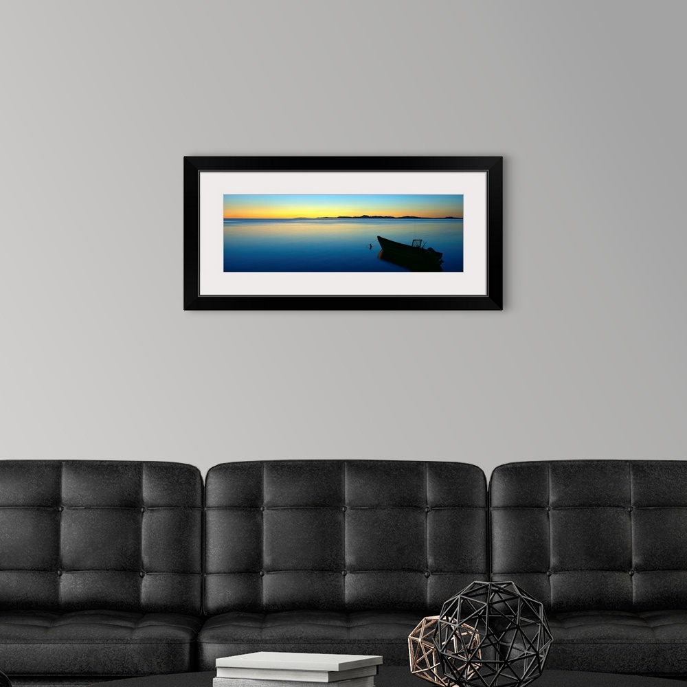 A modern room featuring Lonely boat resting on the calm ocean as the fading sun emits a golden glow on the horizon.