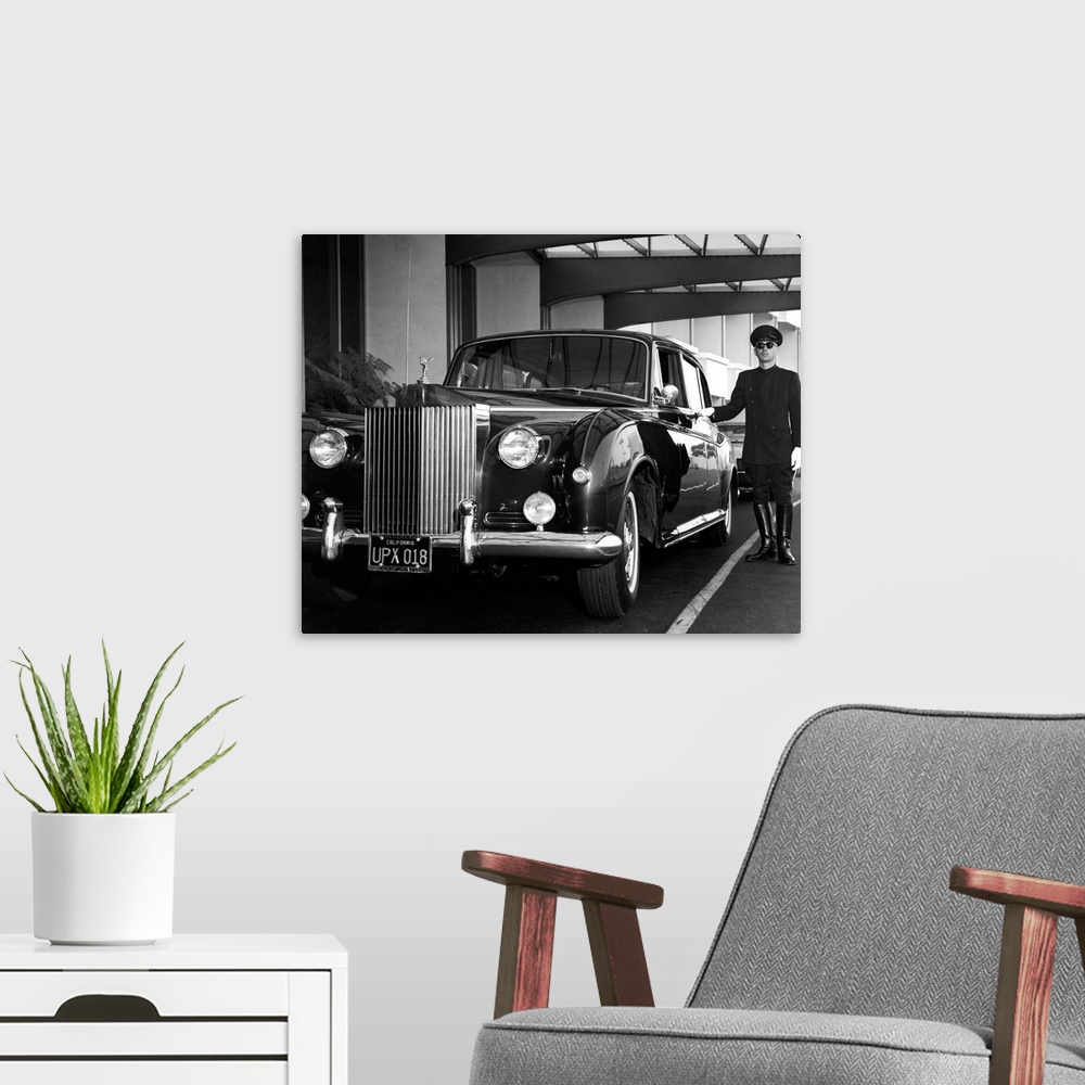 A modern room featuring A uniformed chauffeur waits beside a Rolls-Royce Phantom V, California, circa 1959