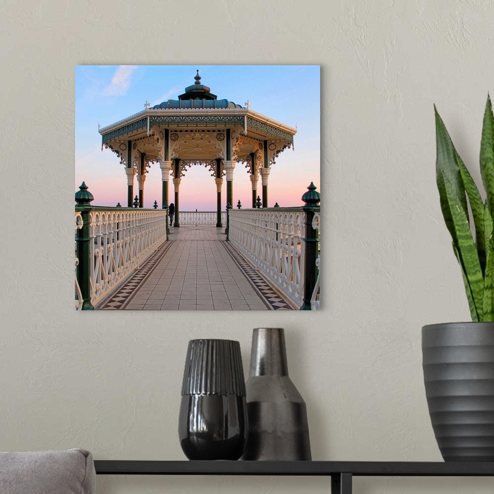 A modern room featuring A lone figure and an almost symmetrical shot of an ornate seaside bandstand. The sky is awash wit...