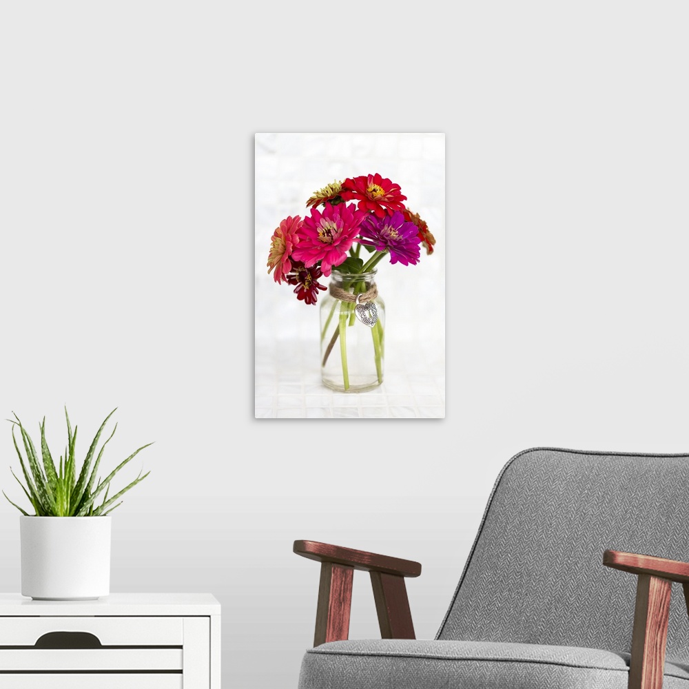 A modern room featuring A variety of colored Zinnia flowers in a simple glass vase with a decorative heart pendant held i...