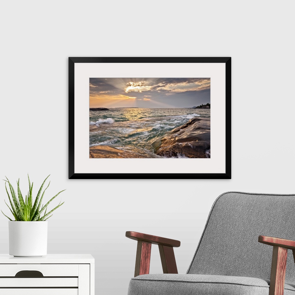 A modern room featuring Burst of sun rays peeking out behind a cloud as waves lap on to a rocky shore.