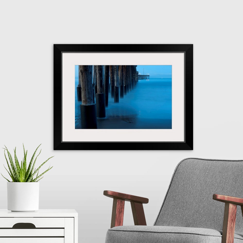 A modern room featuring Large print of tall wooden pier pillars holding up a pier leading into the ocean from the shore.