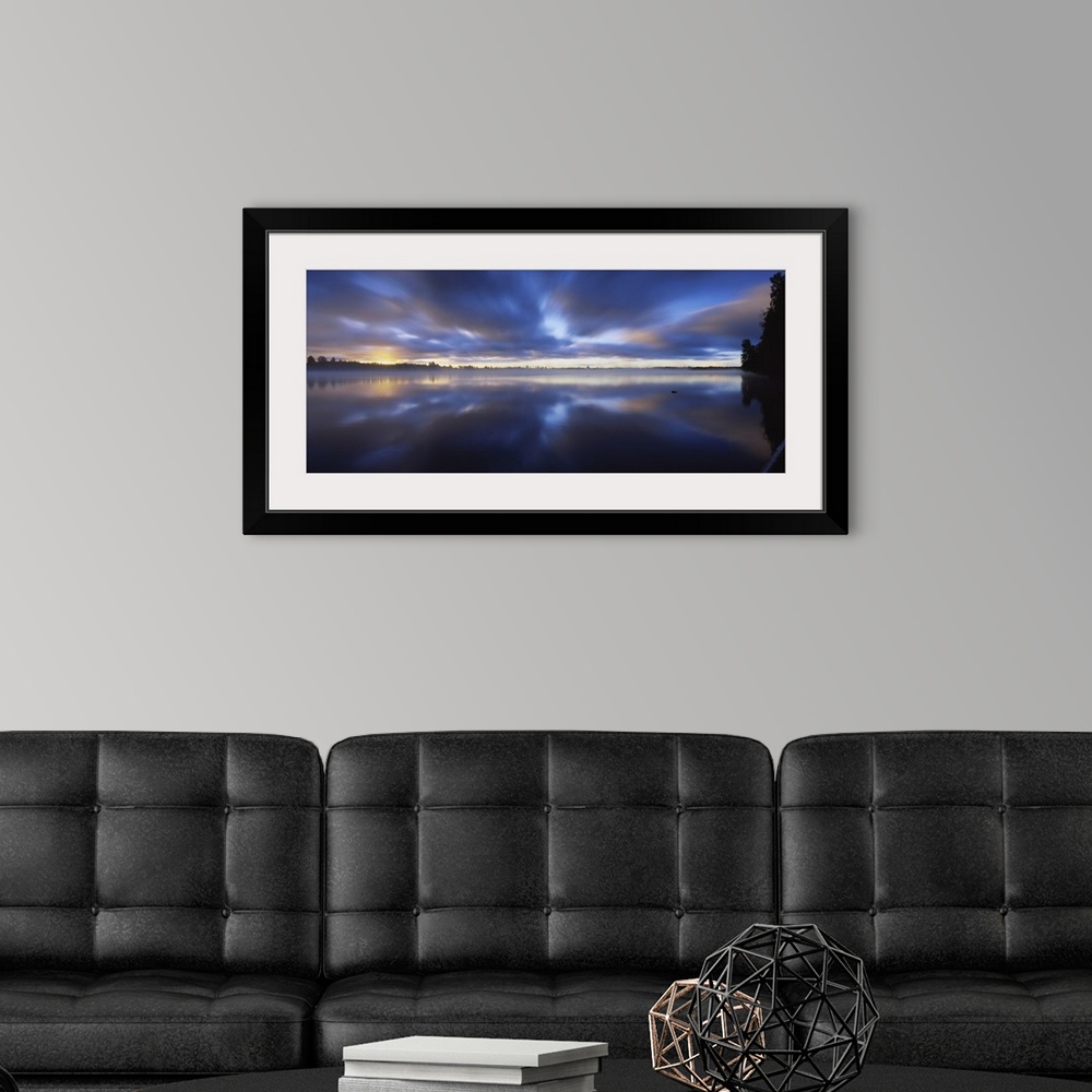 A modern room featuring Panoramic photograph of huge body of water with trees in the distance at sunset.  The sky is clou...