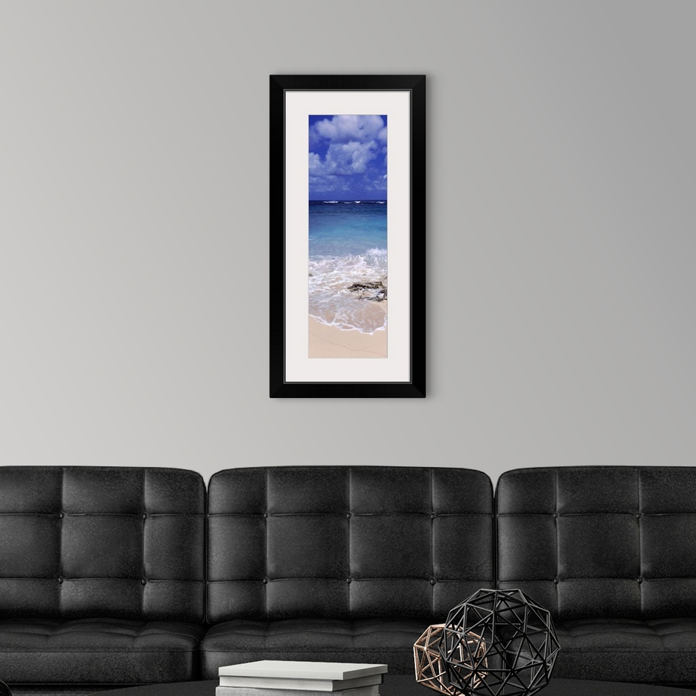 A modern room featuring Vertical, large photograph of clear blue waters hitting the beach beneath a blue sky with billowi...