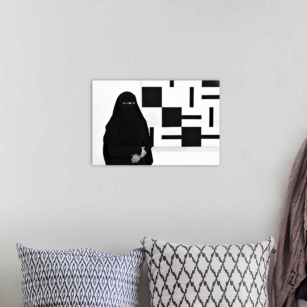 A bohemian room featuring A woman wearing a black niqab stands next to a painting made of black and white squares.