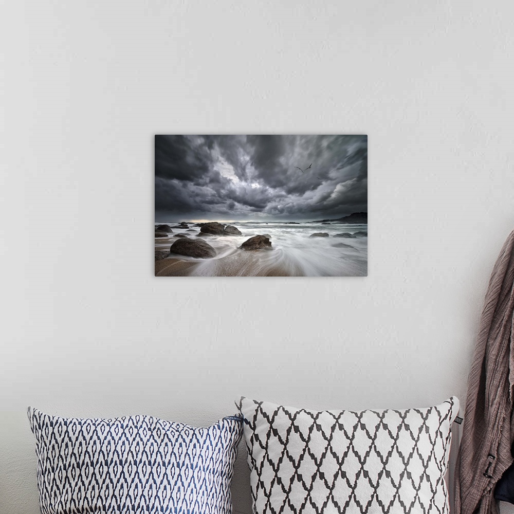 A bohemian room featuring Dramatic clouds hang over a rocky beach with the tide rushing out to sea.
