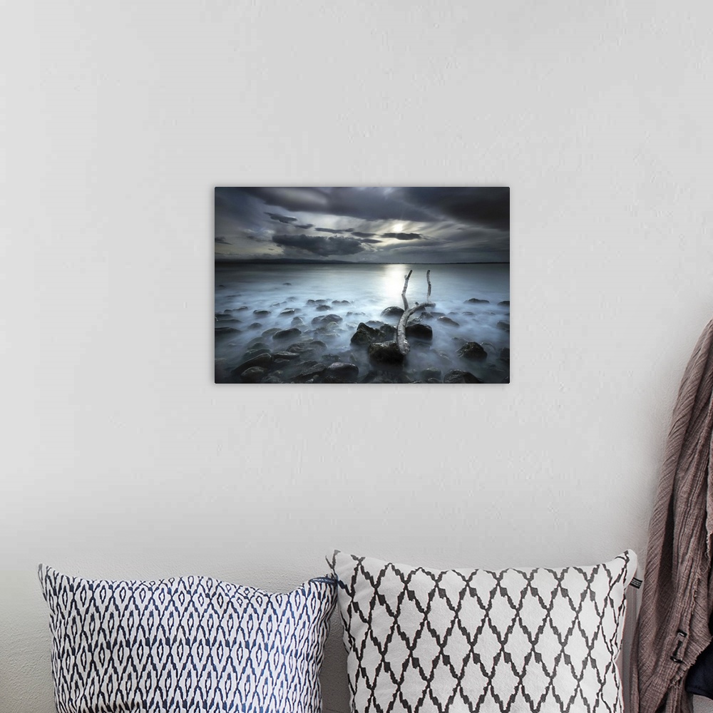 A bohemian room featuring Sun shining through dark clouds onto a misty lake, seen from the rocky shore.