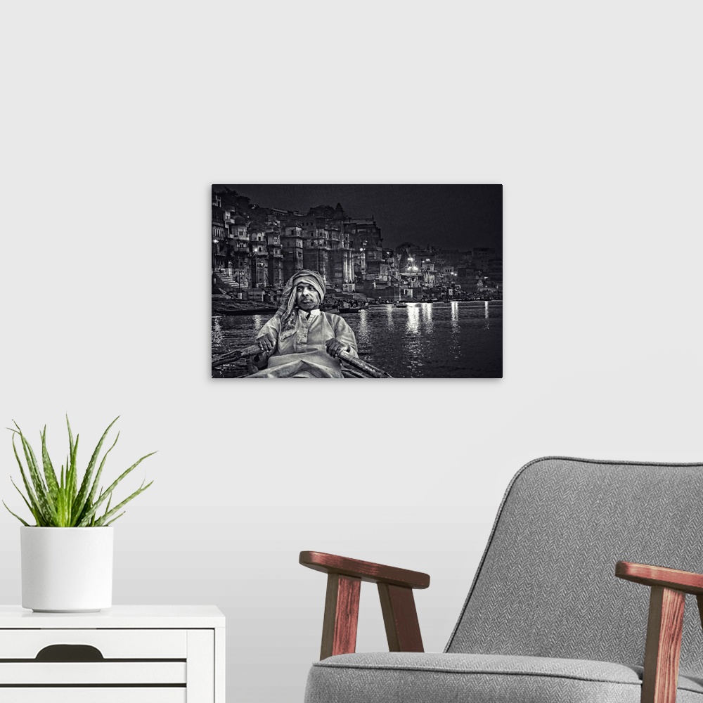 A modern room featuring A man rows a boat on the river near an urban cityscape at night, India.