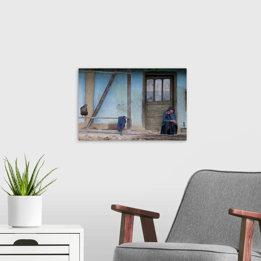 A modern room featuring Portrait of an old woman sitting on the doorstep of a blue house looking at a chicken.